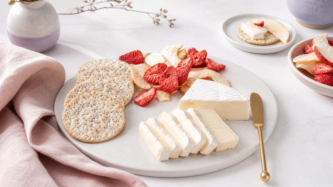 Editorial lunch plate with crackers, sliced soft cheese, freeze-dried strawberries and apples, and pale pink linen on a bright white table