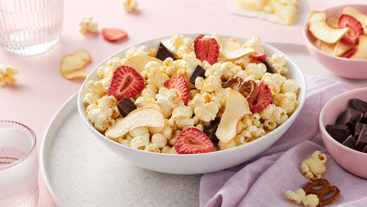 Movie-night snack tray with white bowls of popcorn, freeze-dried strawberries and apples, dark chocolate pieces, and pale pink lavender styling
