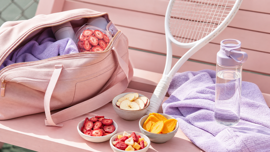 Open tennis bag with a blush towel, racket, water bottle, and bowls of freeze-dried strawberries, apples, and mango on a soft pink bench