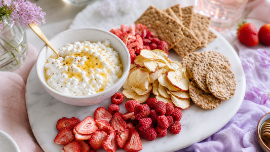 A white marble snack board with cottage cheese, honey, freeze-dried strawberries, raspberries, apples, crispbread, and seeded crackers