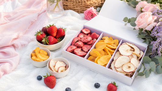 A pastel picnic snack box with freeze-dried strawberries, mango, crackers, cheese, and fresh fruit on a white blanket with blush and lavender styling