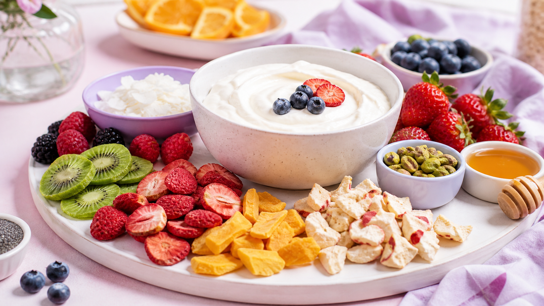 Brunch yogurt toppings board with a large white bowl of yogurt, freeze-dried strawberries and mango, coconut flakes, pistachios, and fresh fruit on a blush and lavender table