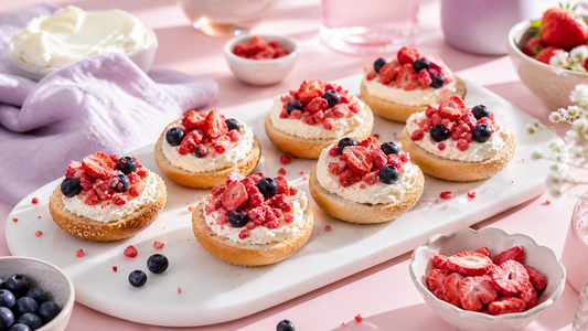 Mini bagel bites topped with cream cheese and freeze-dried strawberries on a white brunch board with blush and lavender styling