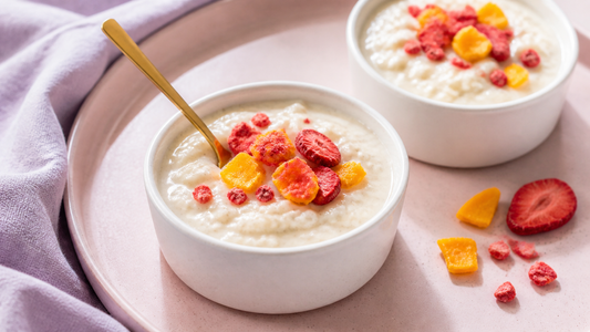 Two white ceramic cups of creamy rice pudding topped with freeze-dried strawberries and mango on a blush brunch tray