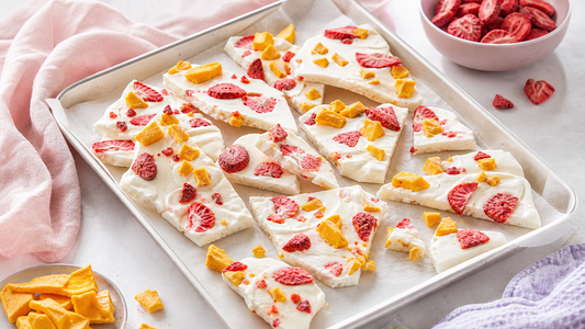 Frozen skyr bark broken into shards on a parchment-lined tray with freeze-dried strawberries and mango on a blush breakfast table