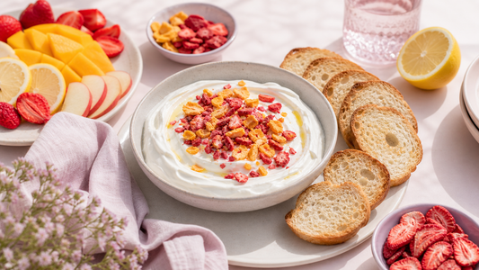 Editorial brunch scene with a lemon yogurt dip bowl topped with freeze-dried strawberries and mango, surrounded by fruit and toast on a pale pink table