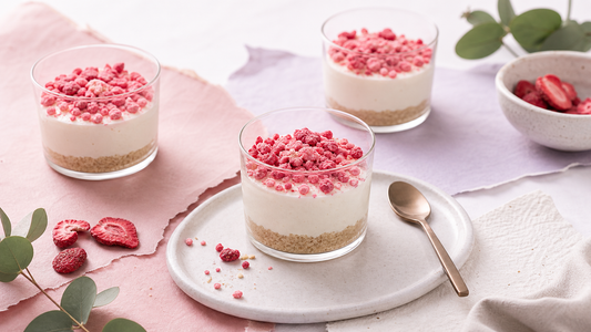 Three no-bake cheesecake cups topped with pink freeze-dried fruit on a white tray with airy blush and lavender styling