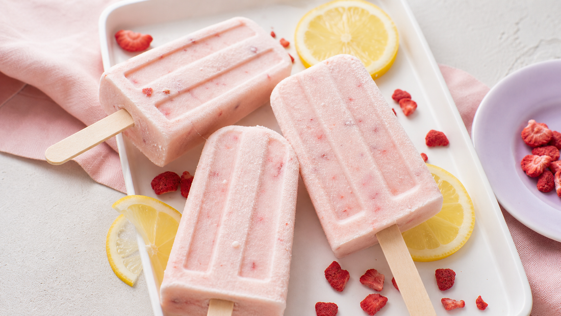 Three pale pink yogurt pops on a white tray with lemon slices, crushed freeze-dried strawberries, and blush linen in soft daylight