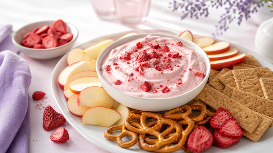 A bowl of strawberry yogurt dip topped with freeze-dried strawberries beside pretzels, apple slices, and graham crackers on a blush table