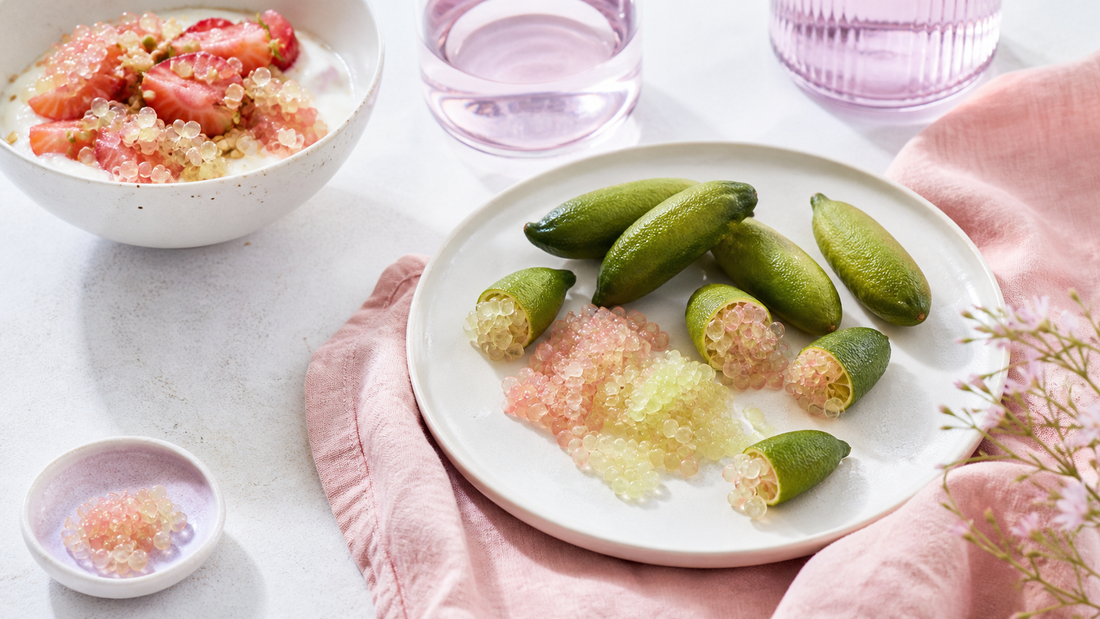 Cut finger limes spilling citrus pearls onto a white plate beside yogurt and soft pink linen with pale lavender styling in bright natural light