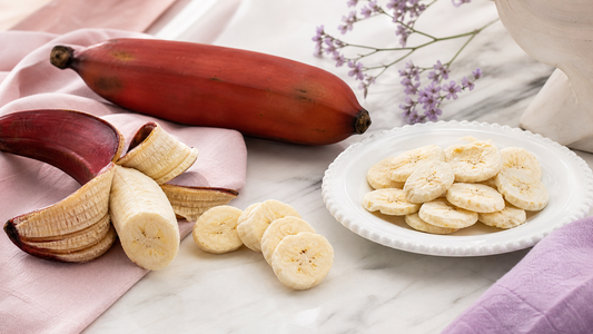 Red banana tasting scene with a peeled red banana, banana slices, and a white plate of freeze-dried banana chips on blush and lavender linens
